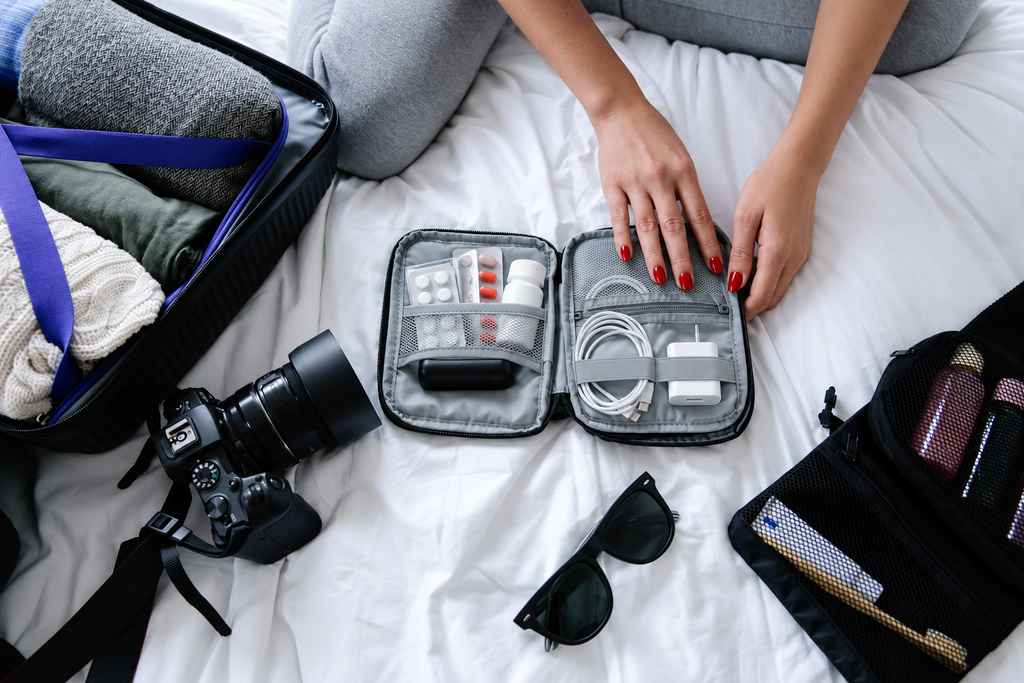 Open suitcase with folded clothes, a camera, sunglasses, a phone charger and travel medication arranged on a bed.